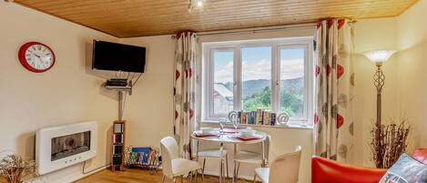 A cozy living room with a wooden ceiling, a fireplace, a TV, a dining area with chairs, a red sofa, and a window with a view of the hills.