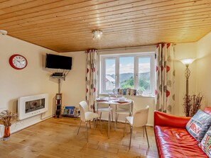 Uma sala de estar acolhedora com teto de madeira, lareira, televisão, área de jantar com cadeiras, sofá vermelho e janela com vista para as colinas.