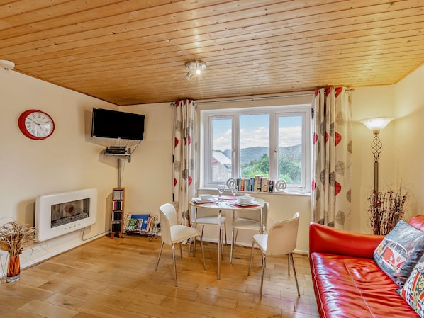 Una sala de estar acogedora con techo de madera, chimenea, televisor, zona de comedor con sillas, sofá rojo y ventana con vista a las colinas.