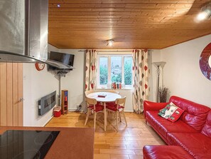 Ein gemütlicher Wohnbereich mit einem roten Sofa, einem Esstisch mit Stühlen, einem an der Wand montierten Fernseher und einem Fenster mit Vorhängen.