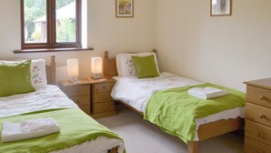 A bedroom with two single beds, each with a green blanket and pillow, a wooden dresser, and a window with a view of a building outside.