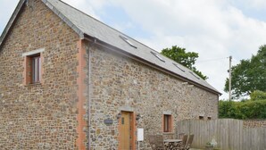 A stone building with a wooden door and windows, a fenced yard, and a table with chairs outside.