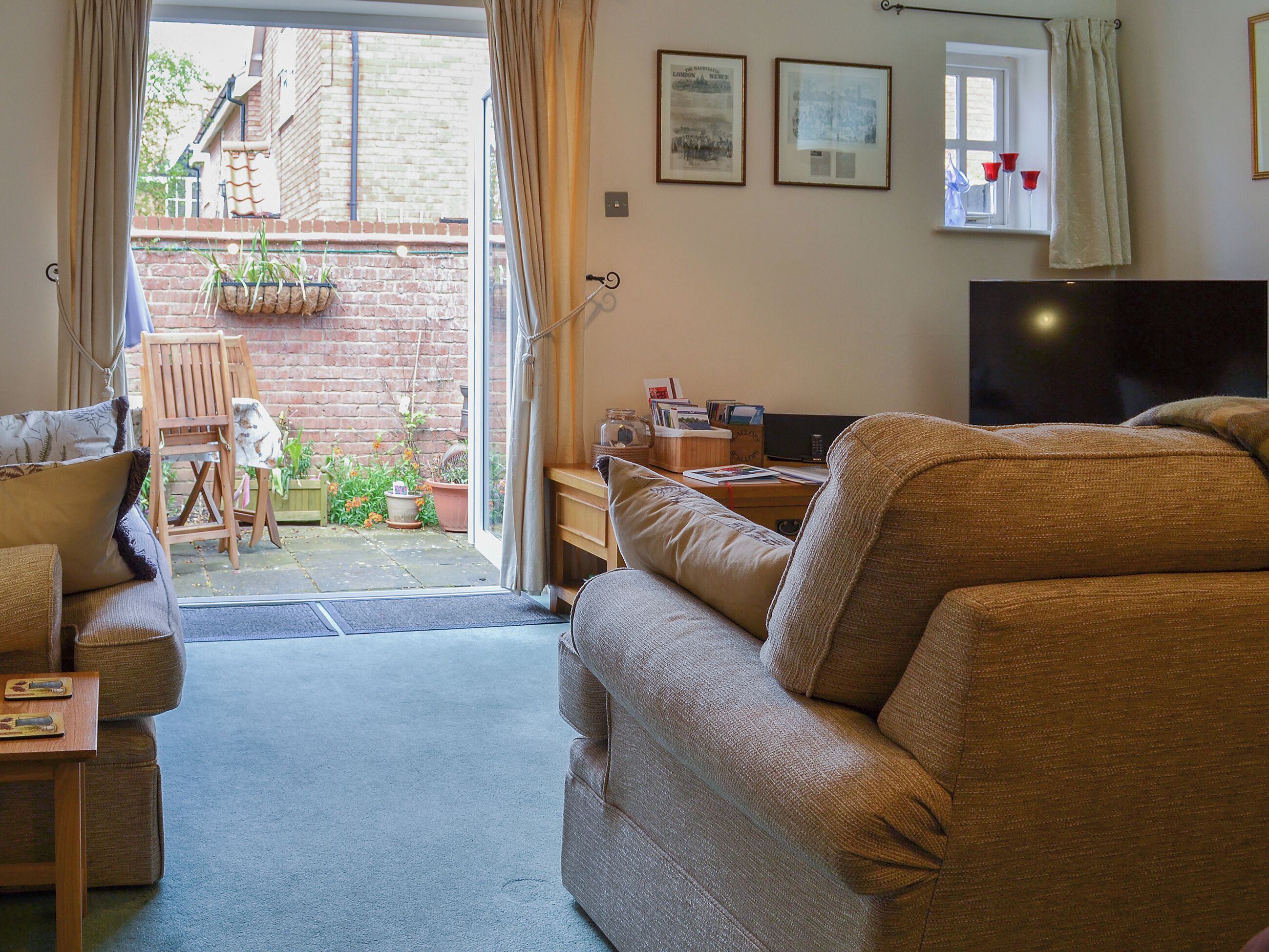 A cozy living room with a sofa, a table, and a TV. There is a window with curtains and a view of a brick wall and plants outside.
