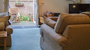 A cozy living room with a sofa, a table, and a TV. There is a window with curtains and a view of a brick wall and plants outside. - 3 bedroom accommodation in Snettisham, near Hunstanton (Snettisham, near Hunstanton)
