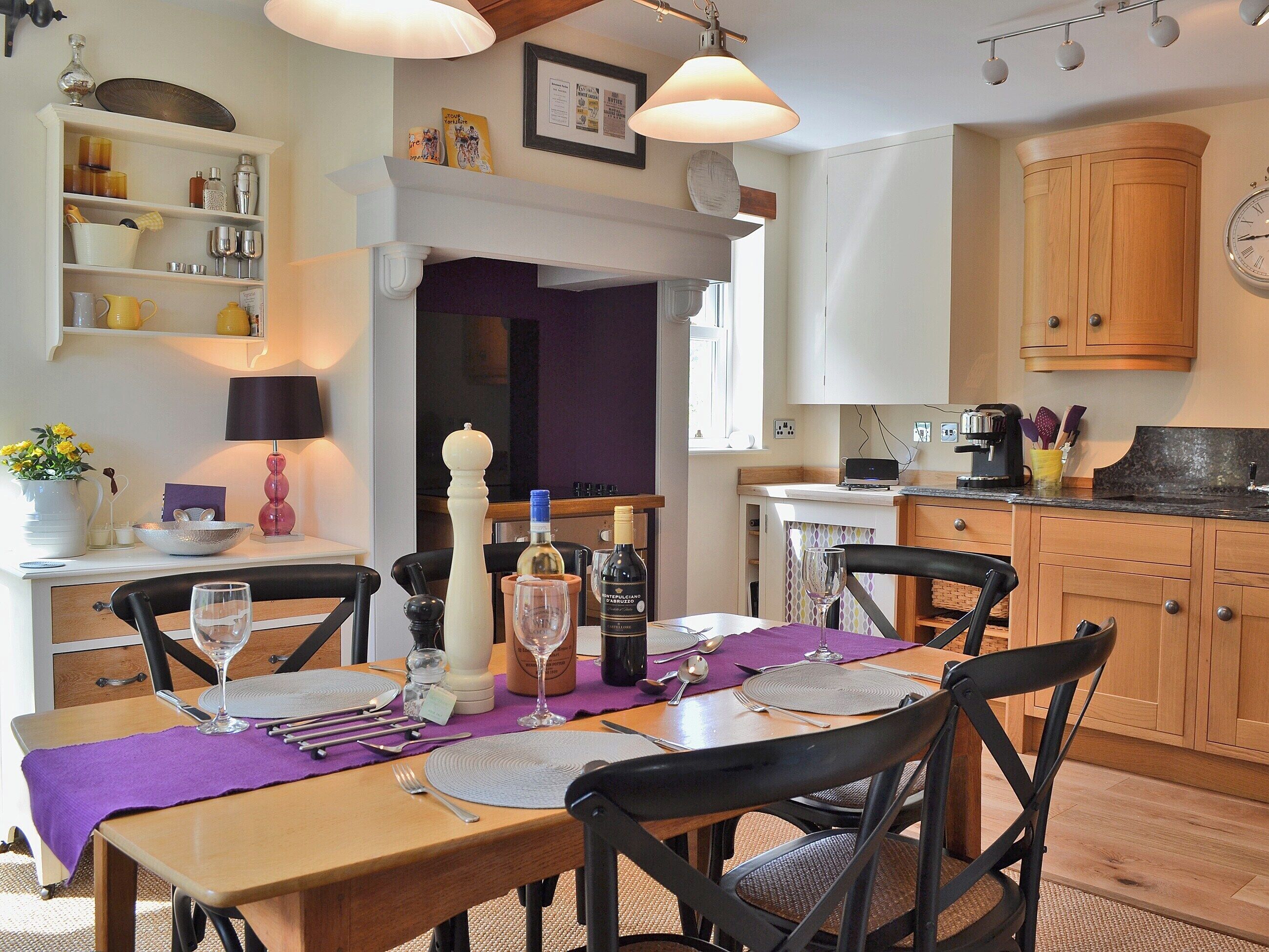 Un comedor con una mesa de madera dispuesta para cuatro personas, rodeada de sillas negras. Se observa también una cocina con armarios de madera y un reloj en la pared.