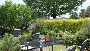 A glass table with four wine glasses and a bottle, surrounded by chairs in a garden with various plants and a large tree. - A pretty stone cottage in the conservation village of Redmire. (Redmire near Leyburn)