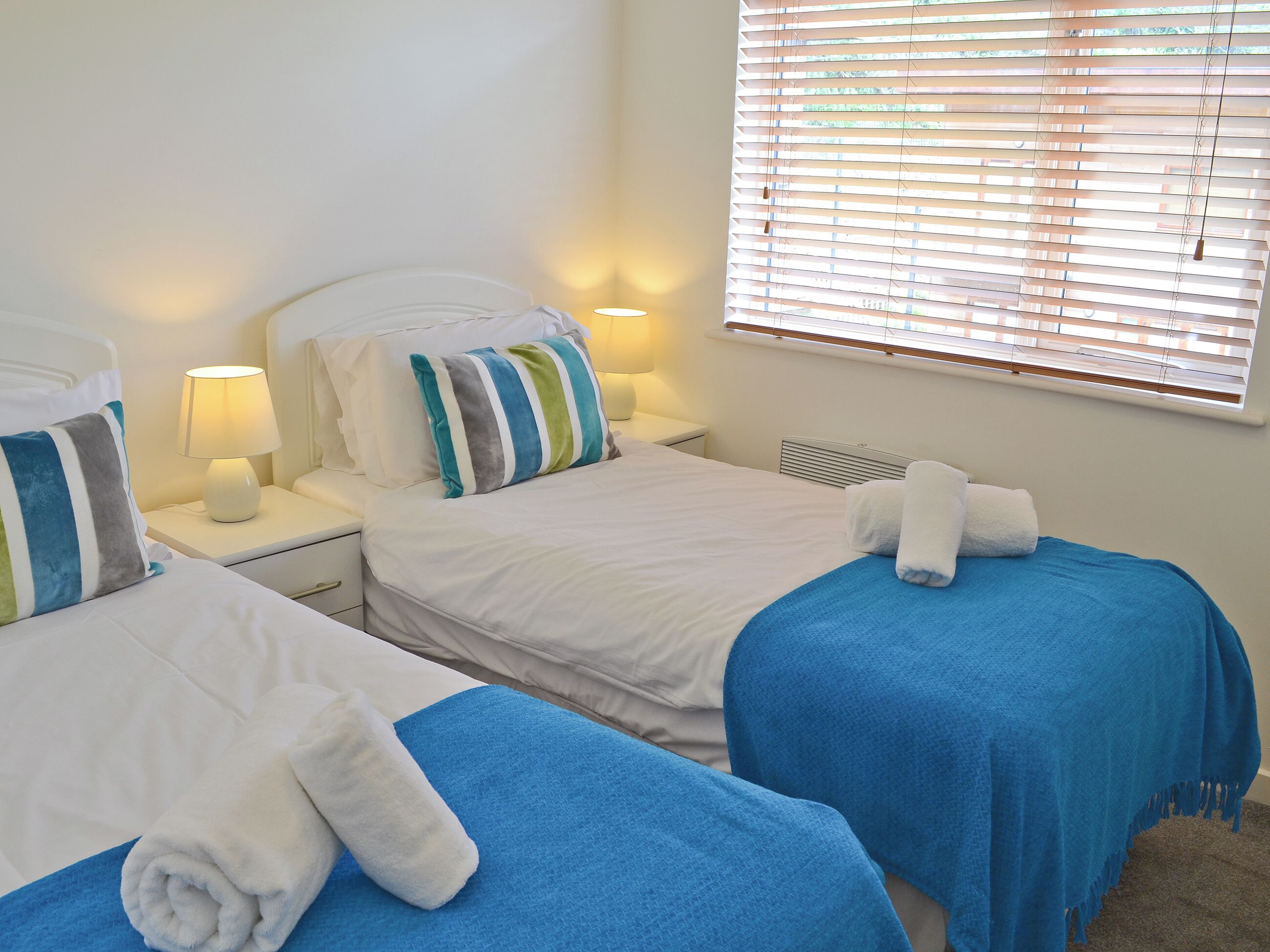 A hotel room with two beds, each with a blue bedspread and white linens. There are two bedside lamps, a window with blinds, and a white wall.