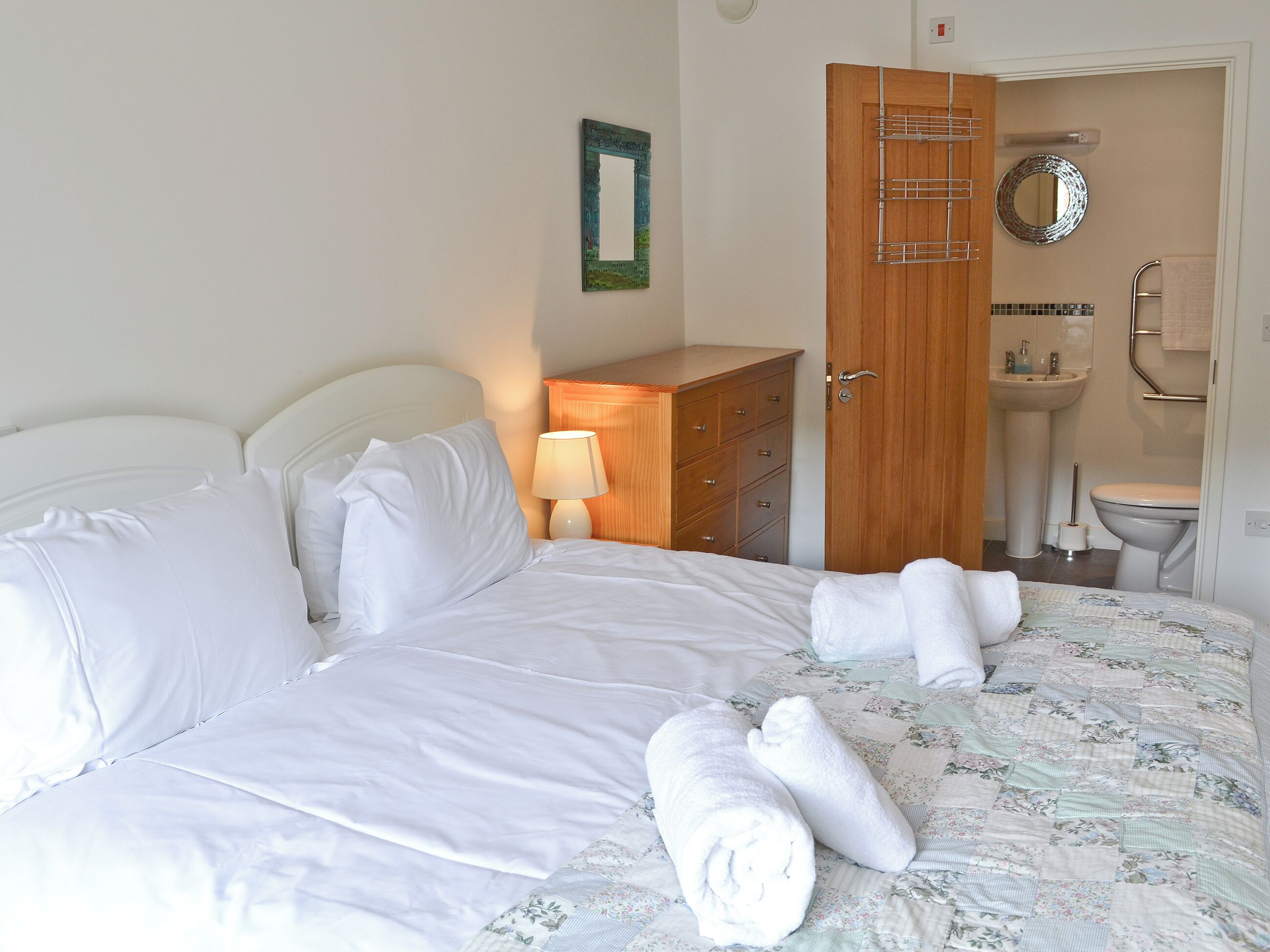 A neatly made bed with white linens, a wooden dresser, a lamp, and a bathroom visible through an open door.