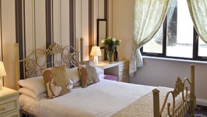 A neatly made bed with a headboard, a bedside table with a lamp, a vase with flowers, and a window with curtains. - Rosecraddoc Manor - Riverview (Near Liskeard)