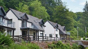 A row of white buildings with grey roofs and balconies, surrounded by lush greenery and a stone bridge. - Rosecraddoc Manor - Riverview (Near Liskeard)
