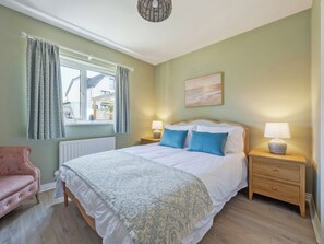 Ein Schlafzimmer mit einem Bett, zwei Nachttischen mit Lampen, einem rosa Sessel, einem Fenster mit Vorhängen und einem Bild an der Wand.