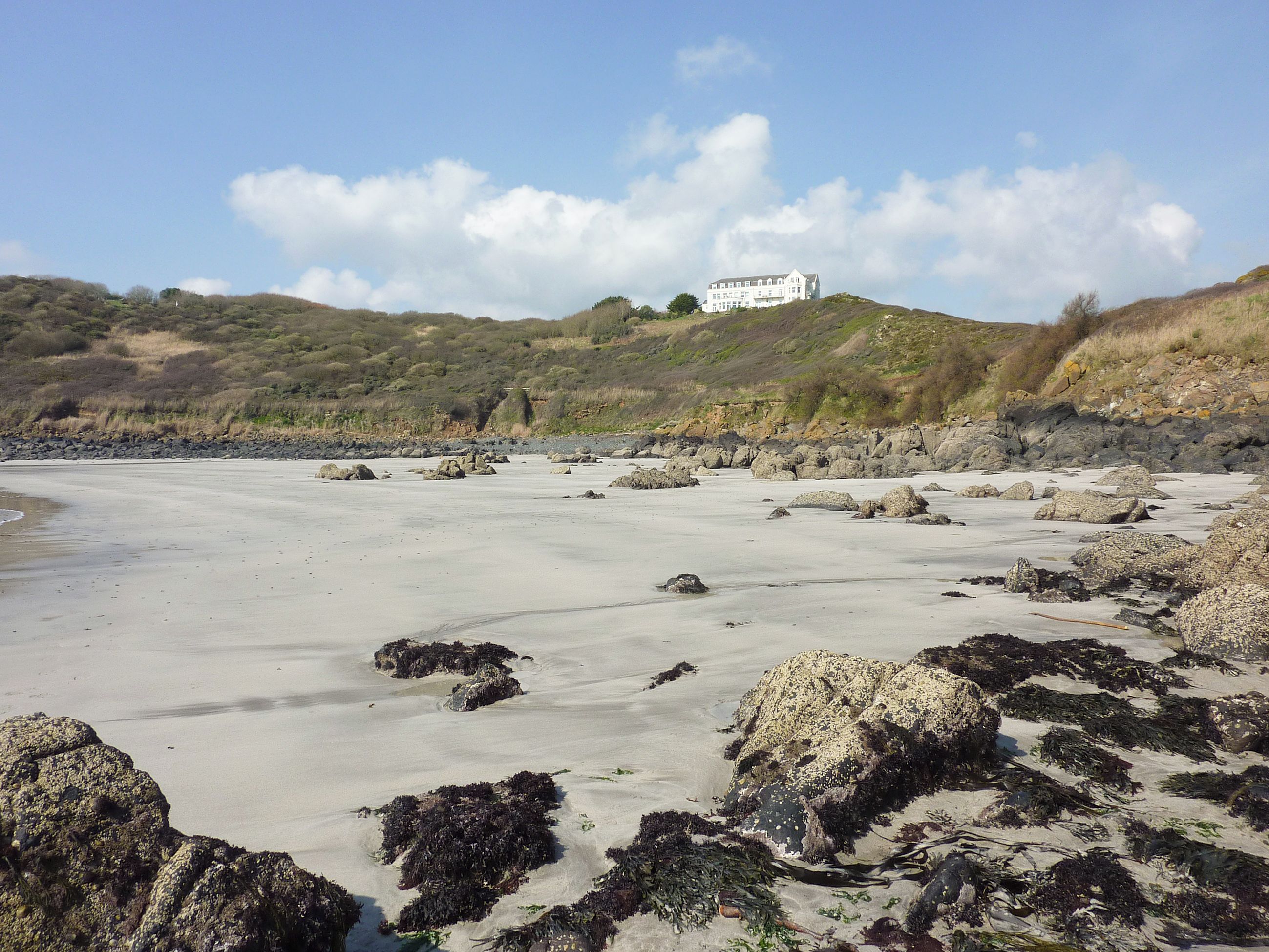 Ein Strand mit Felsen und ein weißes Gebäude auf einem Hügel im Hintergrund.