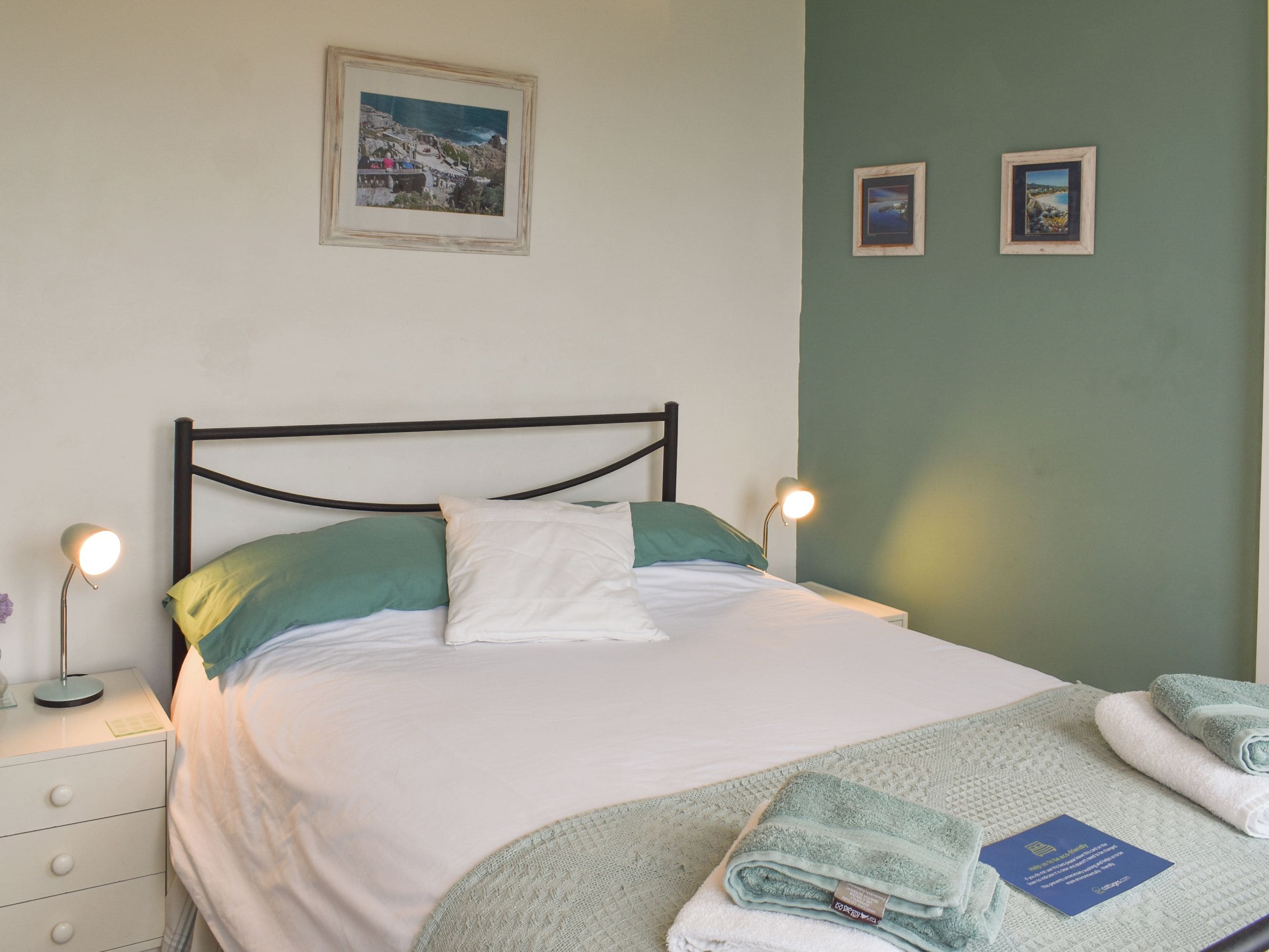 A neatly made bed with pillows, a bedside lamp, a folded towel, and a framed picture on the wall.