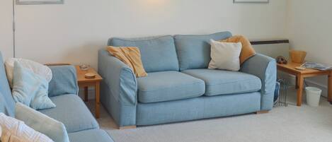 A living room with a blue sofa, a wooden side table, and a floor lamp. There are two framed pictures on the wall.