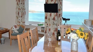A room with a large window offering a view of the sea, a television, a dining table with chairs, and a vase with sunflowers.