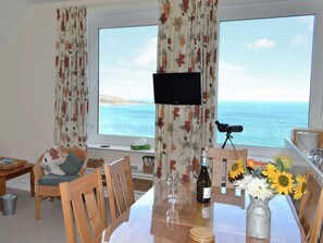 A room with a large window offering a view of the sea, a television, a dining table with chairs, and a vase with sunflowers.