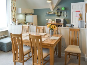 A dining area with a wooden table and chairs, a vase with sunflowers, and a kitchen in the background. - 3 bedroom accommodation in Coverack, near Helston (Coverack, near Helston)