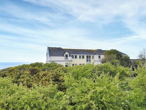 A two-story house with a gabled roof, surrounded by dense greenery and overlooking a body of water. - 3 bedroom accommodation in Coverack, near Helston (Coverack, near Helston)