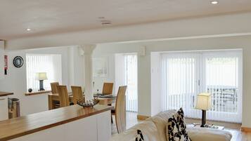 A spacious living area with a sofa, a dining table with chairs, a kitchen counter, and a view through large windows.