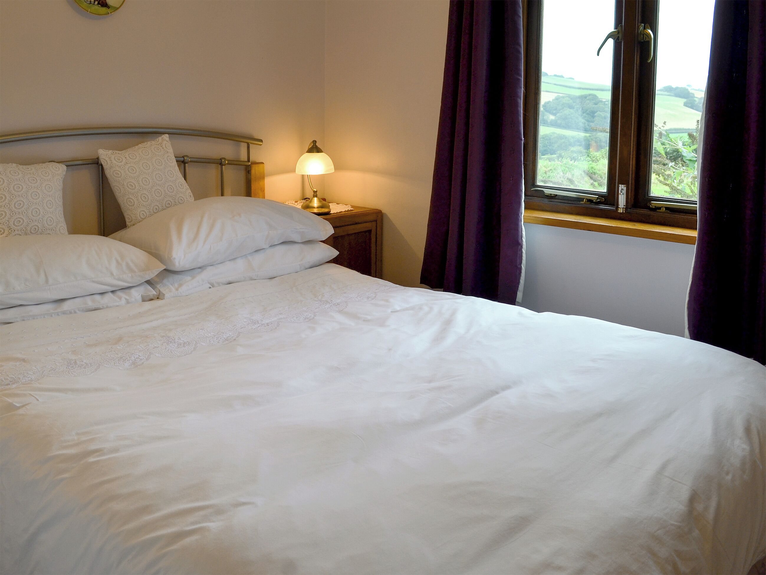 A neatly made bed with white linens and pillows, a bedside lamp, and a window with purple curtains.