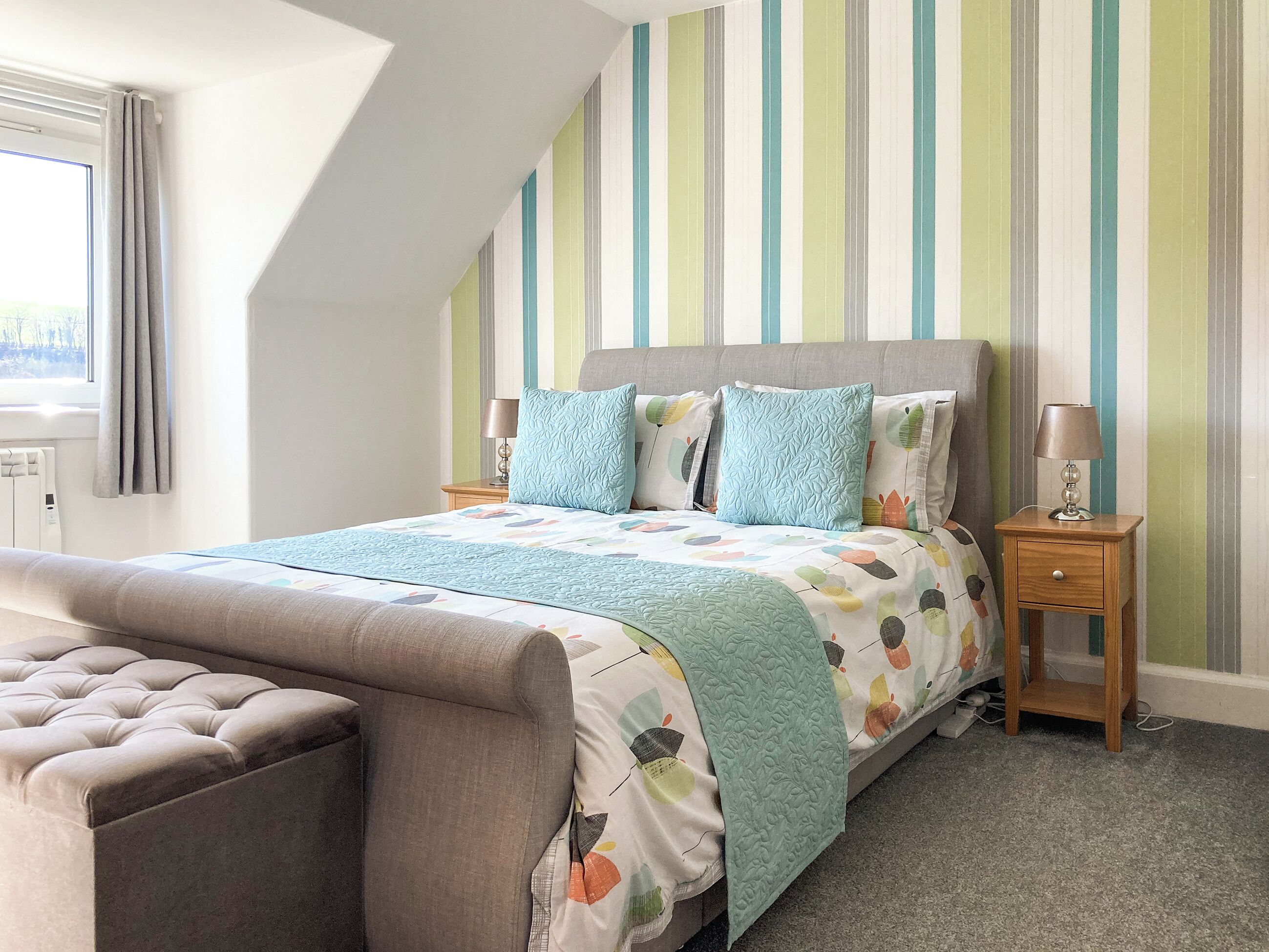 A bedroom with a bed, a nightstand, a lamp, a window with curtains, and a radiator.