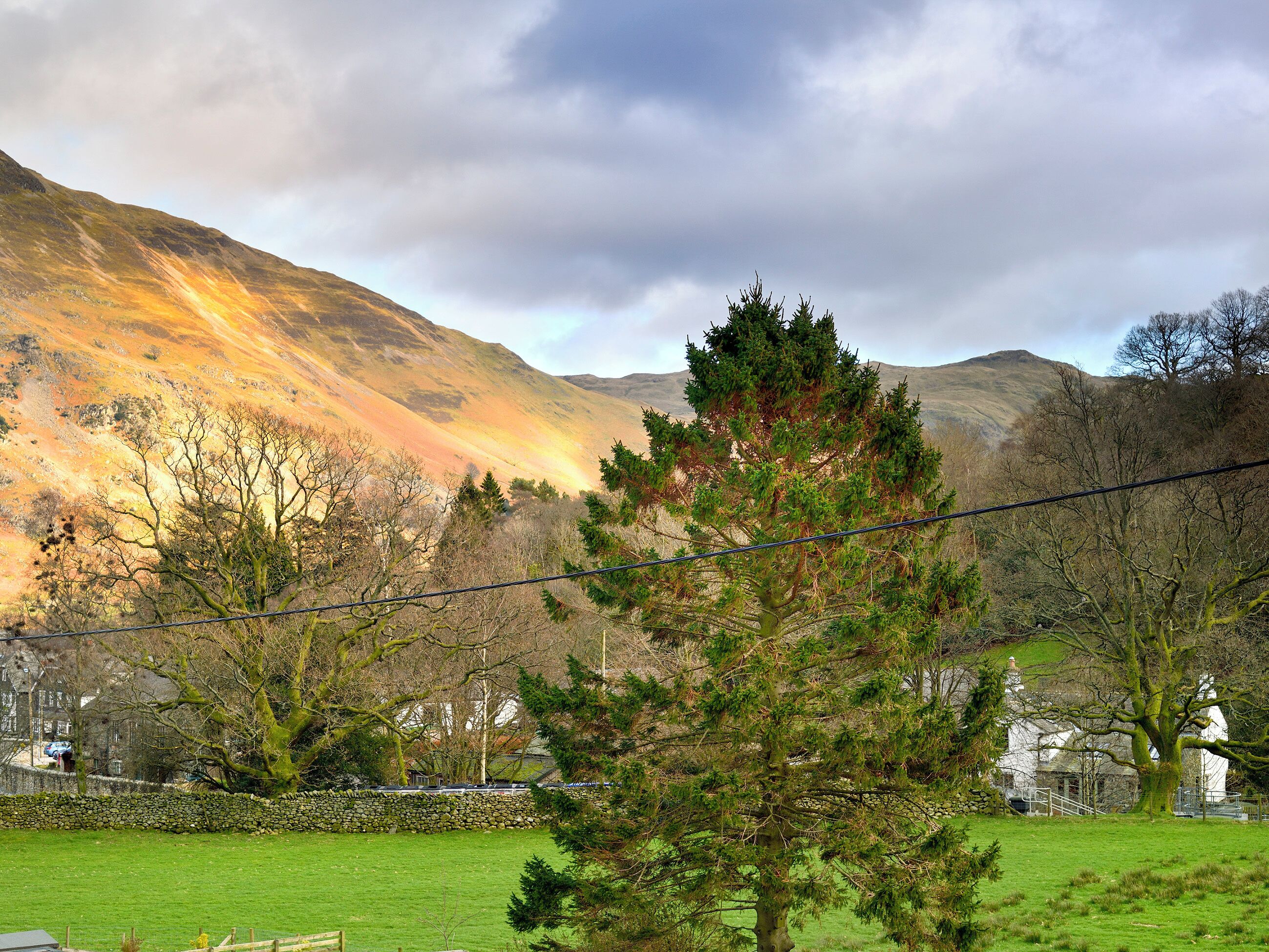 A rural landscape with a stone wall, a grassy field, and a hill with a rainbow in the background.