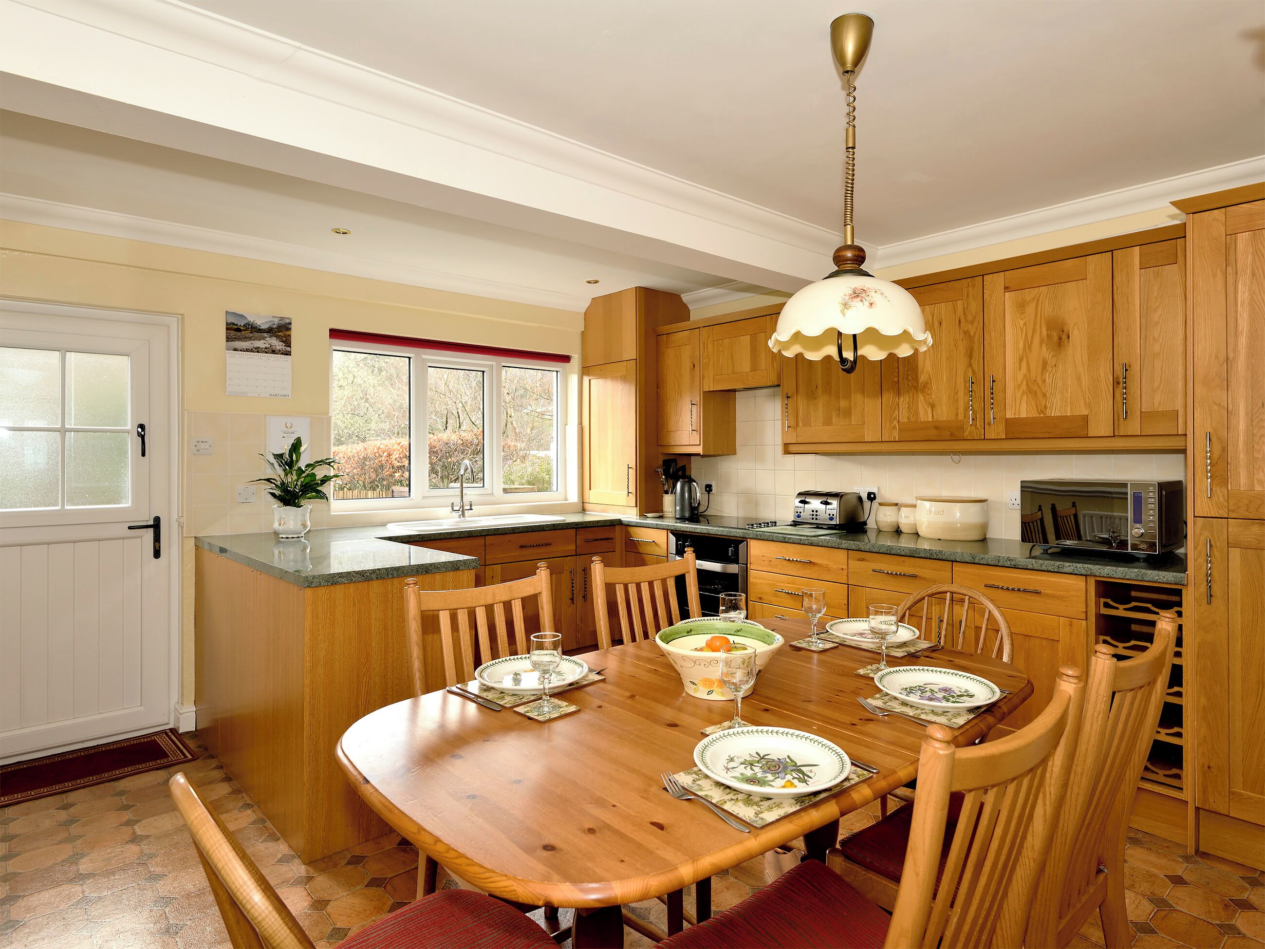 A kitchen with wooden cabinets, a dining table set for four, and a window with a view of trees.