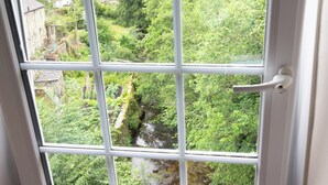 Vista dalla finestra su un paesaggio verdeggiante, con un ruscello, muri in pietra e un edificio in lontananza.