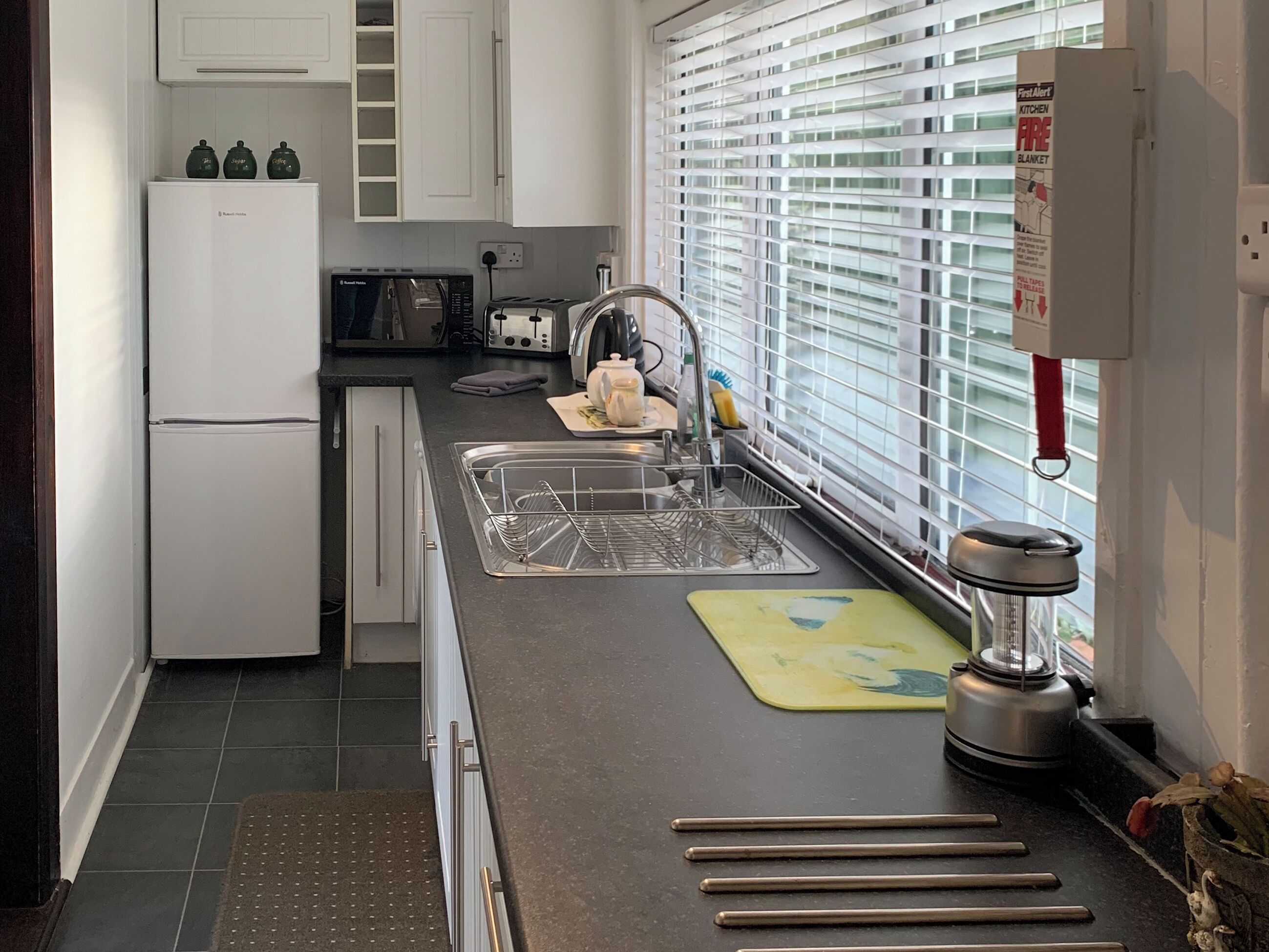 A kitchen with white cabinets, a microwave, a toaster, a sink with a dish rack, and a window with blinds.