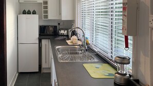 Une cuisine avec des meubles blancs, un four à micro-ondes, un grille-pain, un évier avec un égouttoir et une fenêtre avec des stores.