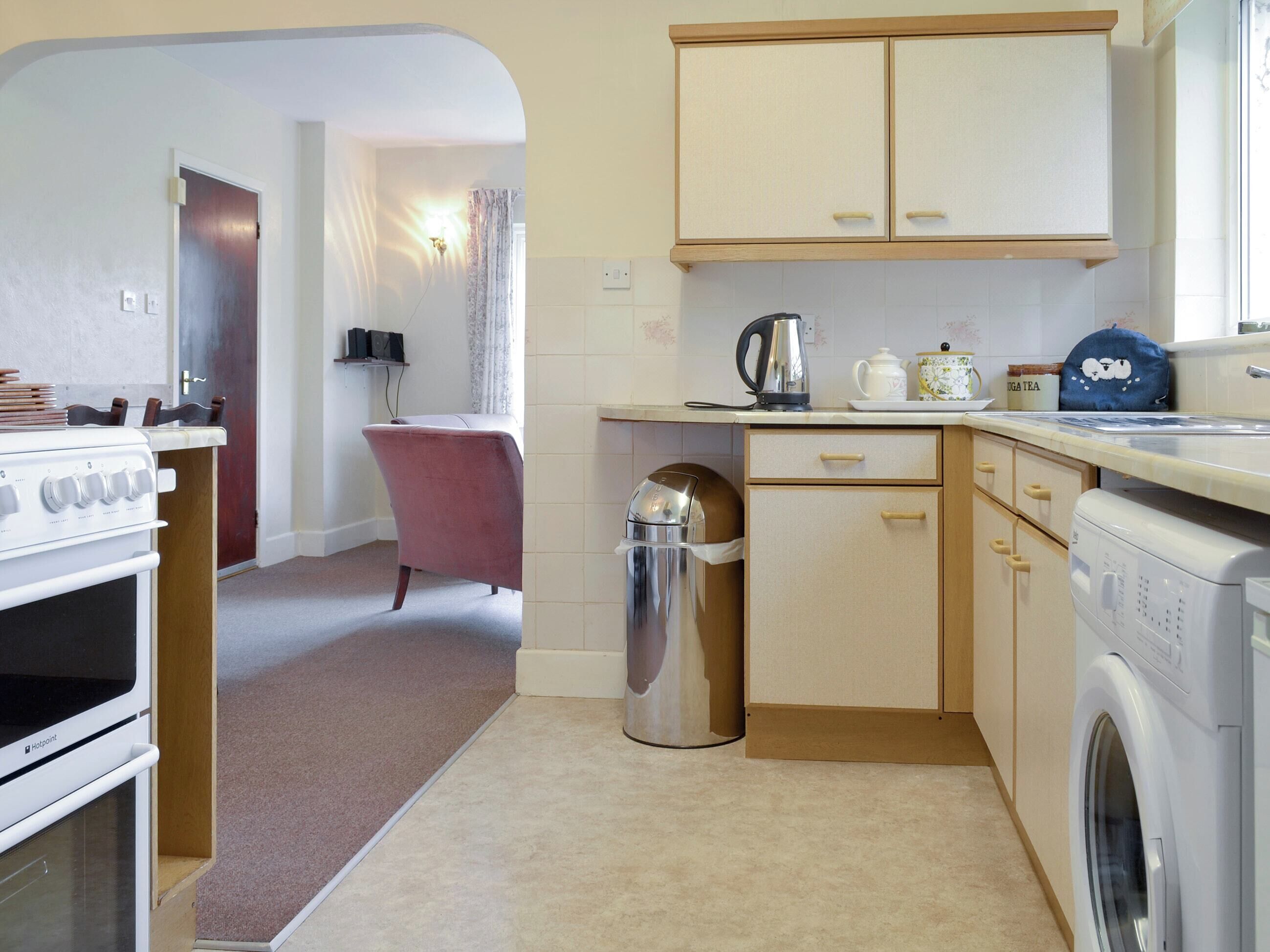 A compact kitchen with a stove, sink, and washing machine. There is a trash can and a chair in the room.