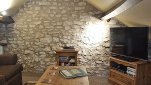 Un accogliente soggiorno con parete in pietra, un tavolo di legno con libri e un telecomando, una televisione su un supporto in legno e un divano marrone.