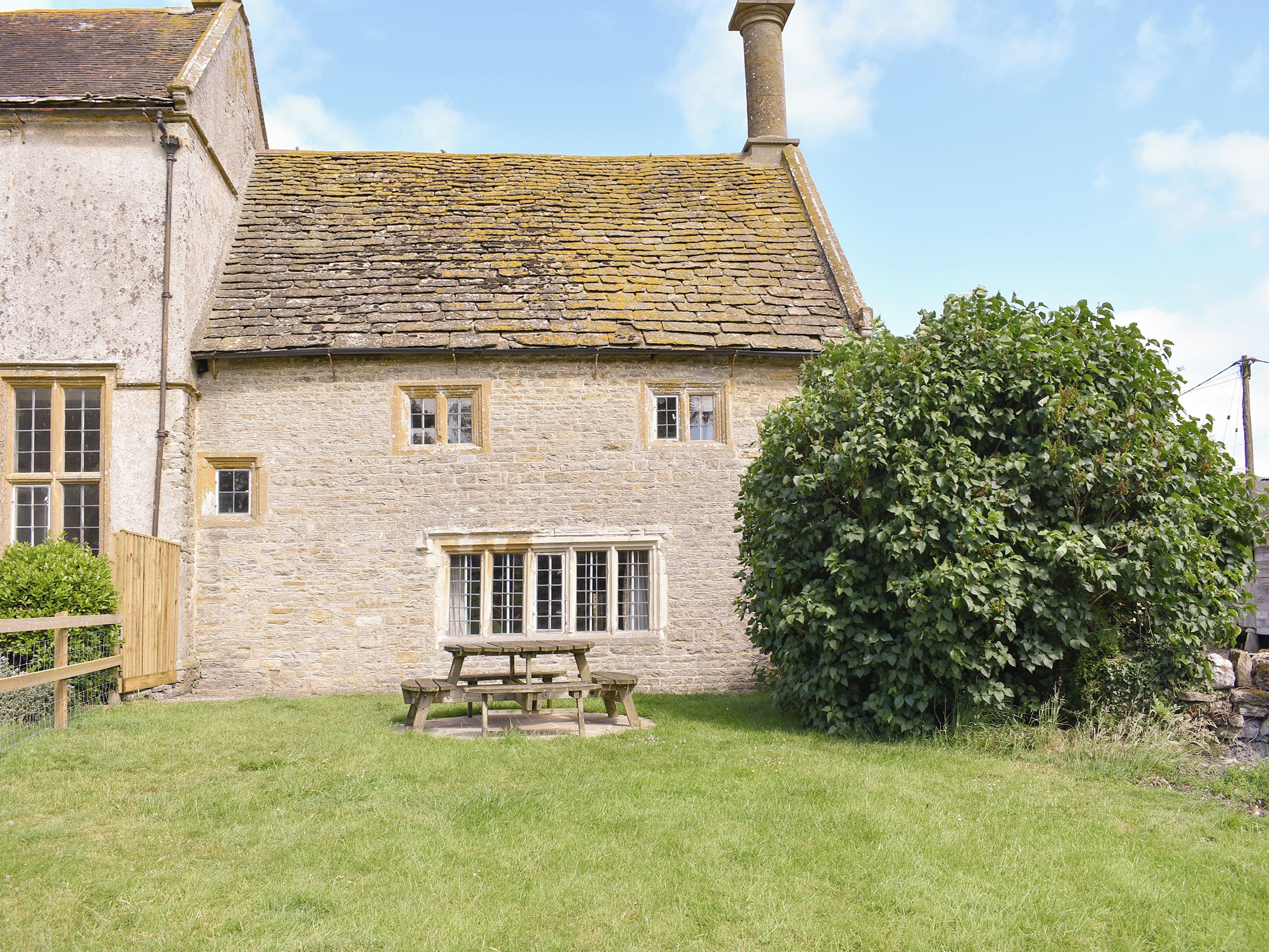 A stone house with a tiled roof, a wooden fence, and a large tree in the yard.