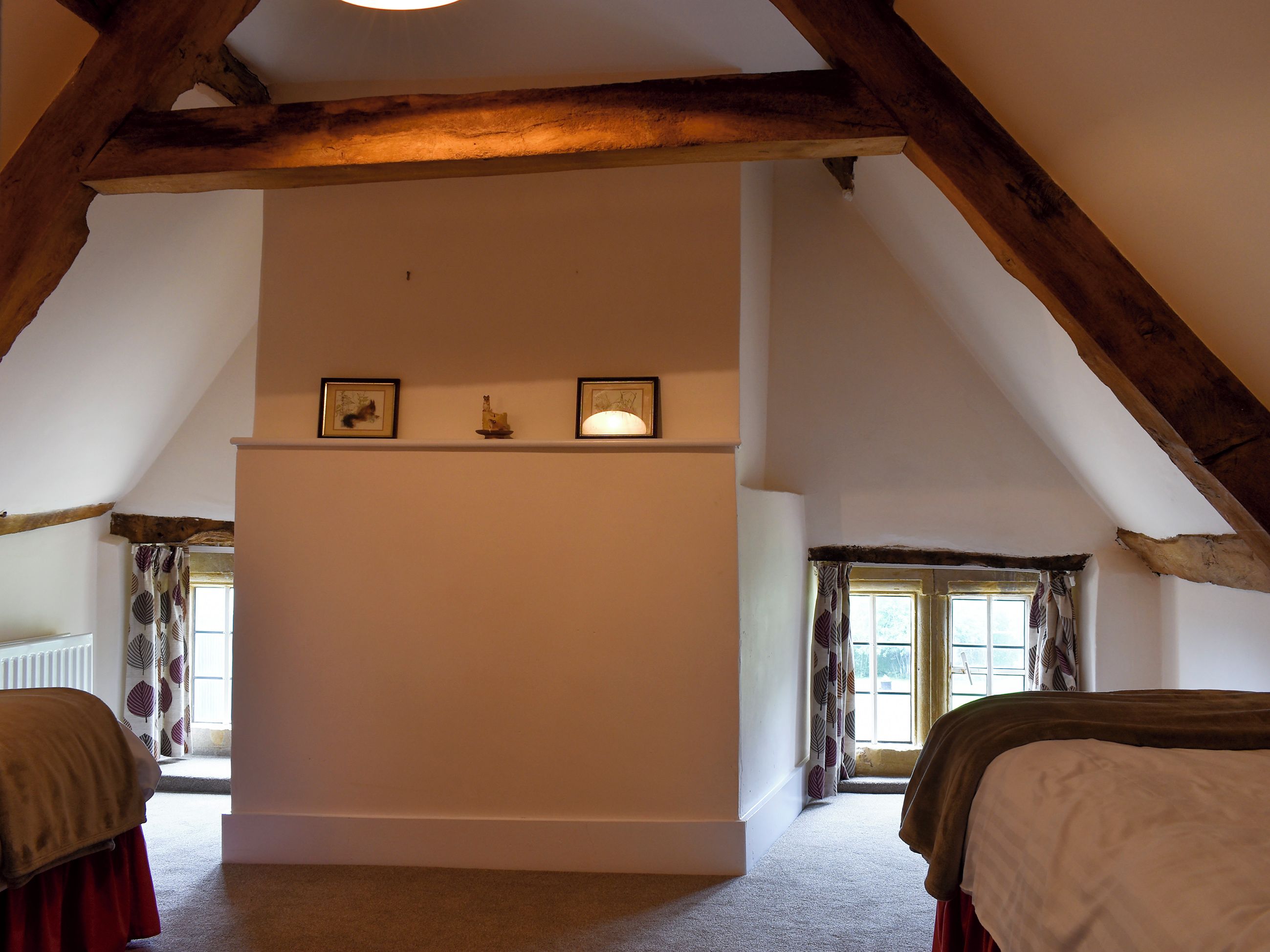 Ein Zimmer mit schrägverdachter Decke, Holzbalken, einer weißen Wand mit Ablagefläche, zwei Fenstern mit Vorhängen und einem Bett mit roter Bettdecke.