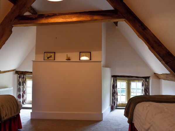 A room with a sloped ceiling, wooden beams, a white wall with a shelf, two windows with curtains, and a bed with a red skirt.
