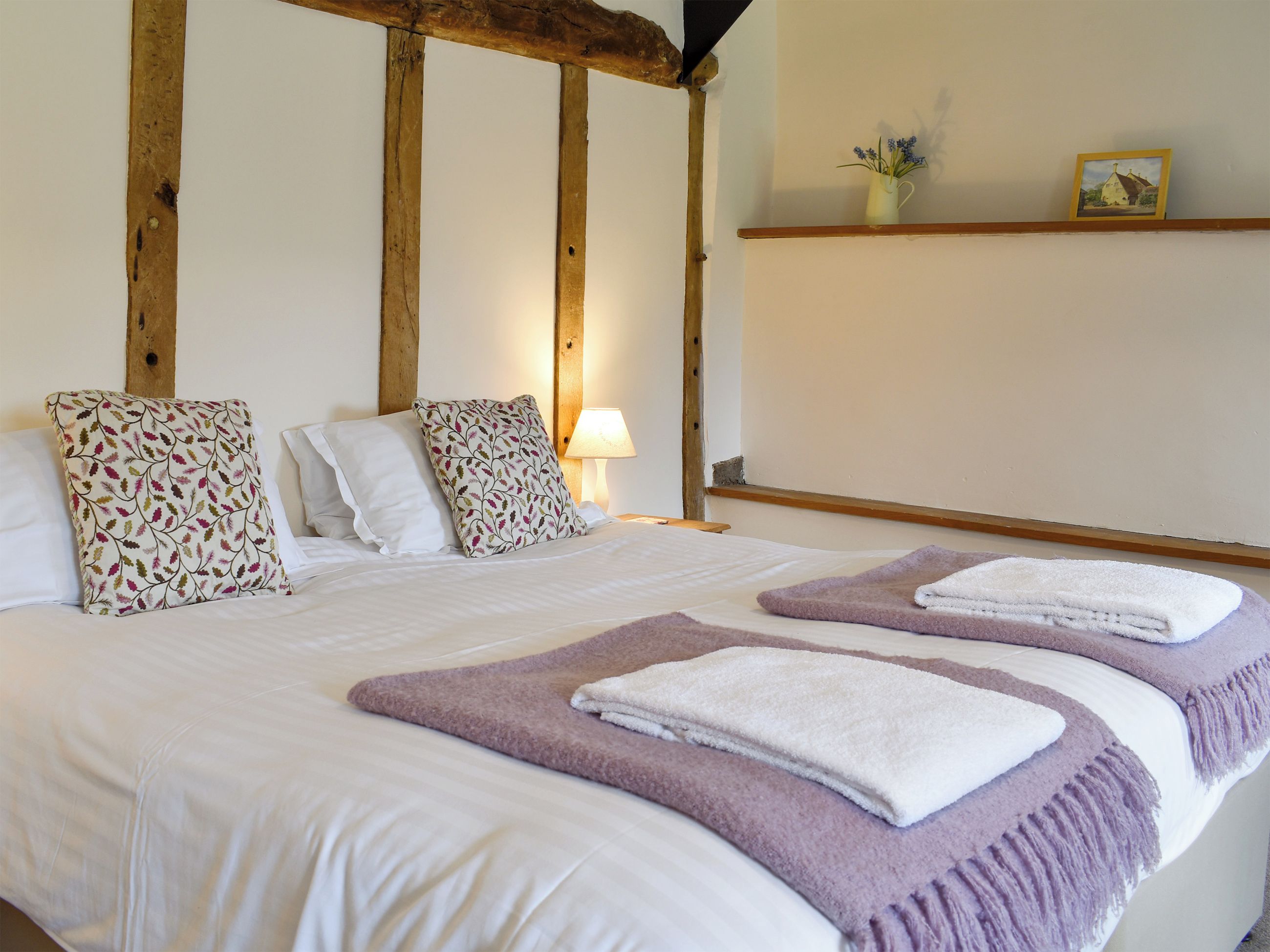 A bed with white linens, two decorative pillows, a bedside lamp, and a shelf with a picture and a vase.