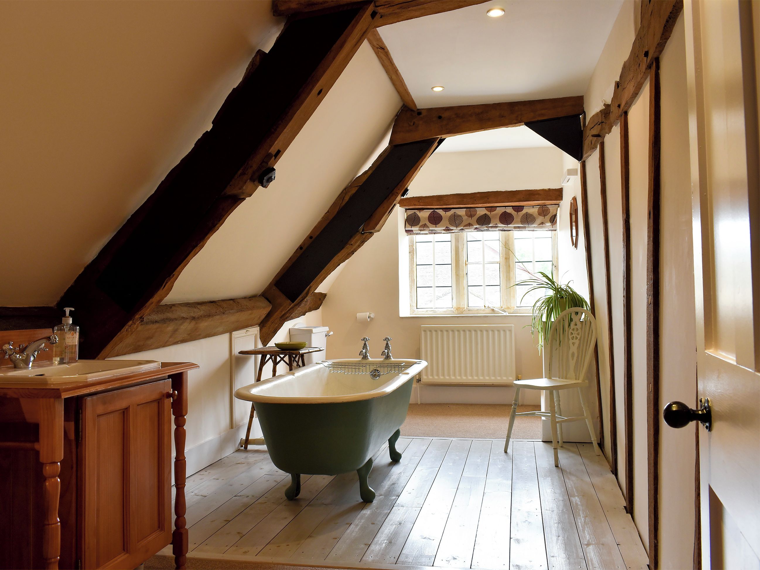 A bathroom with a clawfoot bathtub, wooden beams, a window with a leaf-patterned valance, and a radiator.