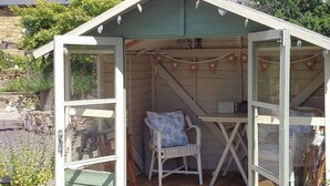 A wooden shed with a table and a chair inside, a potted plant outside, and a stone wall in the background. - Hadrians Garden Villa (Henshaw, near Haltwhistle)