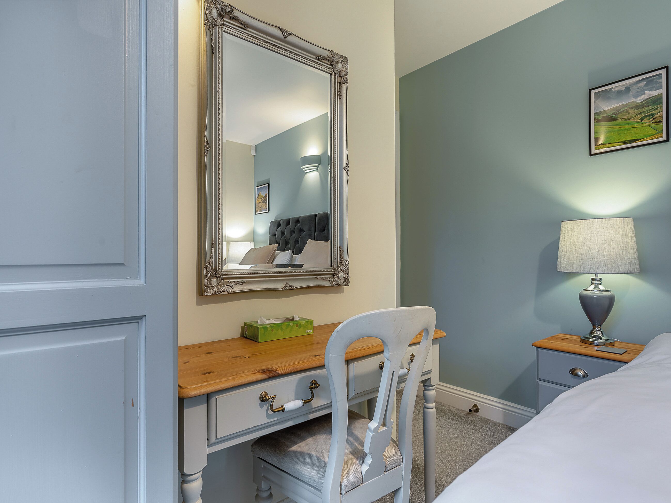 Une chambre à coucher avec une commode en bois, une chaise, une table de chevet avec une lampe et un tableau encadré au mur.