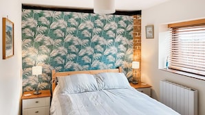 Quarto com cama, duas mesas de cabeceira, um radiador, uma janela com persianas e uma parede com papel de parede estampado com folhas.