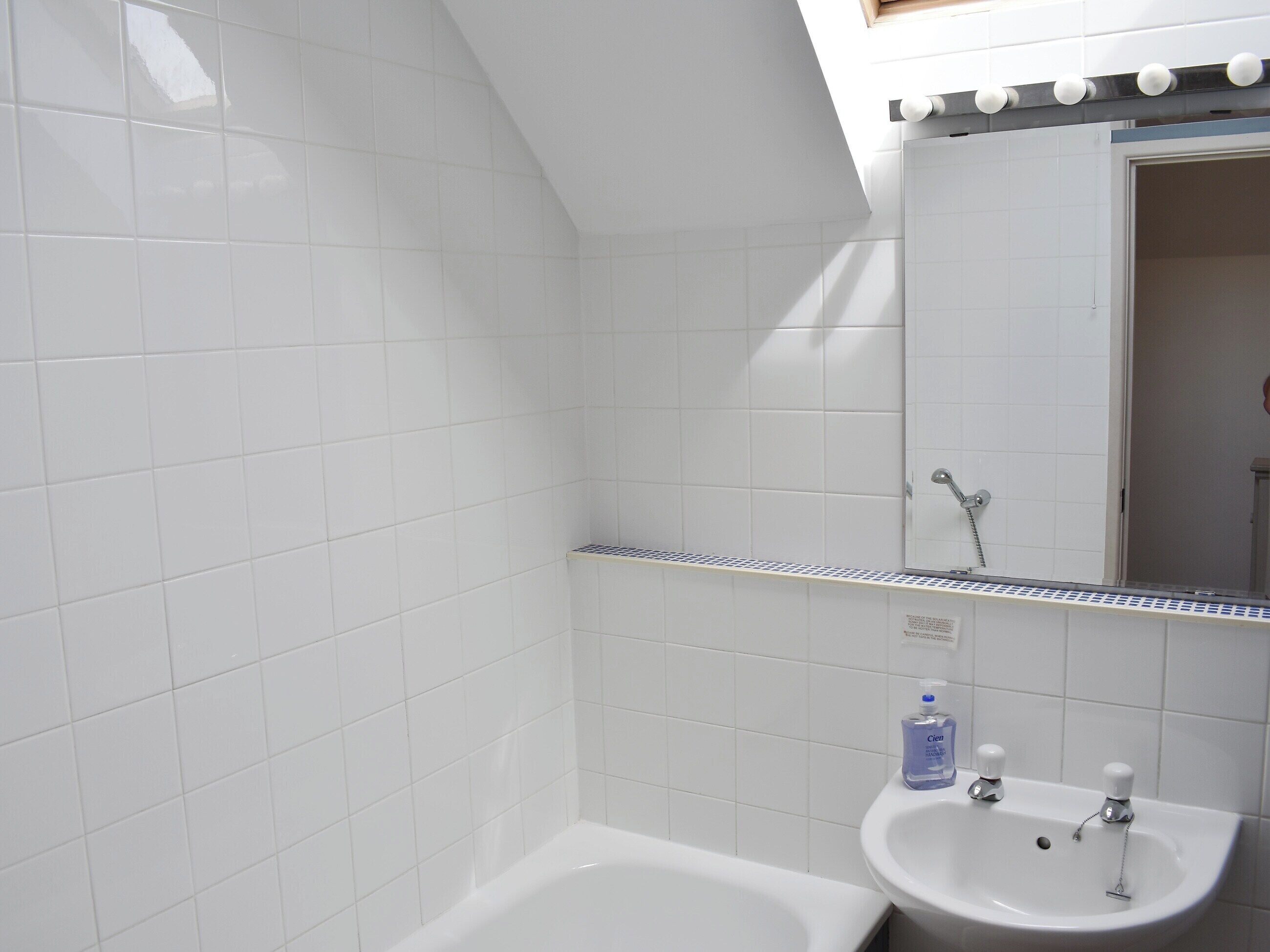 Un baño con azulejos blancos, una bañera, un lavabo con un espejo arriba y un cabezal de ducha.