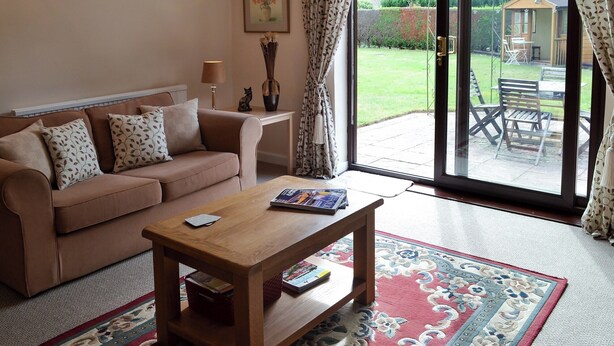 A living room with a sofa, a coffee table, and a rug. There is a window with curtains, a picture on the wall, and a garden visible outside. - 1 bedroom accommodation in Polstead (Polstead)