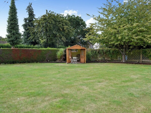 A small wooden shed with a door and windows, surrounded by a grassy yard and a hedge.