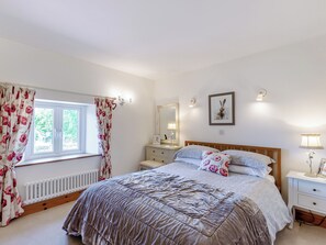A bedroom with a large bed, a nightstand, a dresser, a window with floral curtains, a radiator, and a framed picture on the wall. - New Park Farm - Church Cottage (near Keswick)