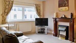 A living room with a leather sofa, a flat-screen TV, a fireplace, and a window with curtains. - 2 bedroom accommodation in Keswick (Keswick)