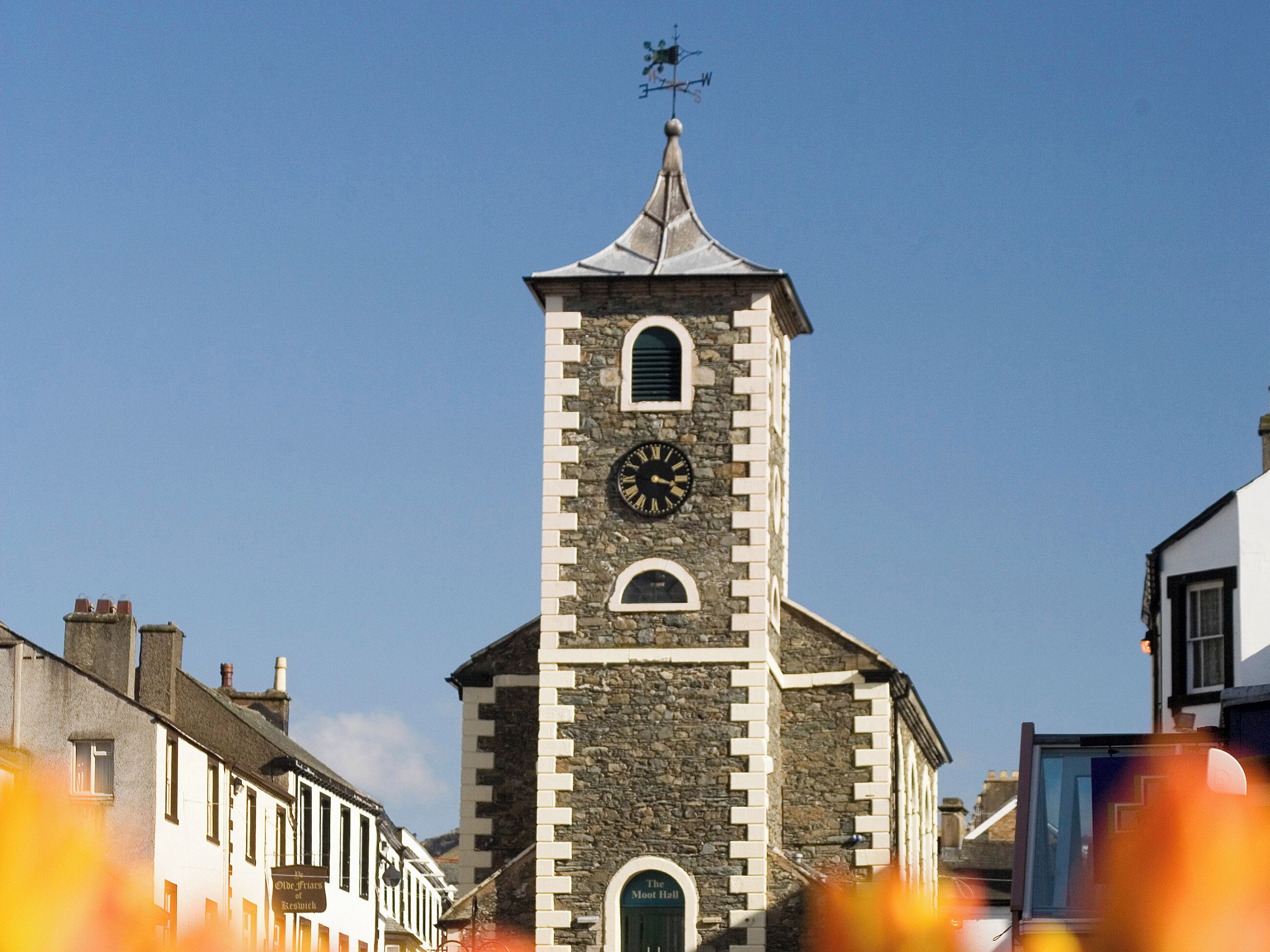 Une tour d’horloge en pierre, surmontée d’une girouette, entourée de bâtiments.
