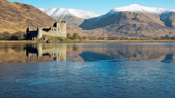 Un castillo histórico junto a un lago tranquilo con montañas nevadas al fondo.