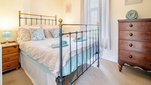 A bedroom with a metal bed frame, a wooden dresser, a nightstand with a lamp, and a window with curtains.
