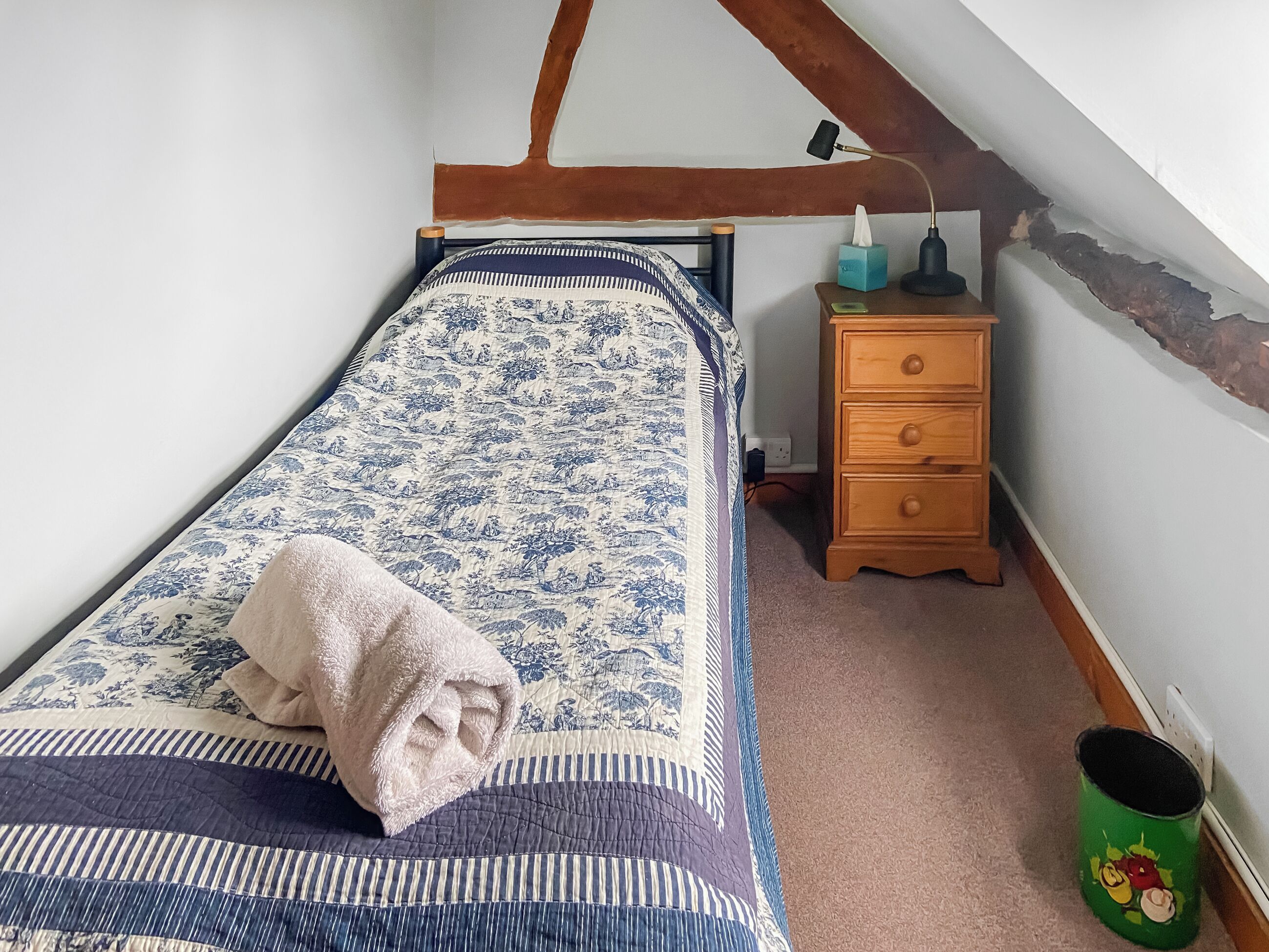 A bedroom with a bed, a nightstand, a lamp, a wooden dresser, and a green trash can.