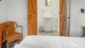 Quarto com cama, cadeira de vime, guarda-roupa de madeira e banheiro visível por uma porta aberta.